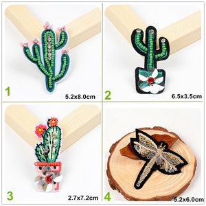 May include: Four embroidered iron-on patches featuring a cactus design with rhinestones and a dragonfly design with gold and black beads. The patches are displayed on wooden surfaces. The measurements of each patch are listed below the image.