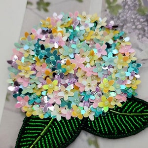 May include: A colorful hydrangea flower brooch made with iridescent sequins. The flower is surrounded by two green leaves, also made with sequins.