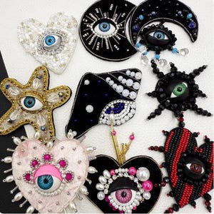 May include: A collection of eight decorative brooches featuring eye motifs. The designs include heart, star, and crescent moon shapes adorned with beads, sequins, and pearls in black, white, gold, pink, and red, each centered with a realistic eye.