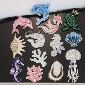 May include: A selection of beaded brooches featuring marine life, such as a blue dolphin, pink seahorse, and green frog with a crown. Other designs include coral and shells, all crafted with colourful beads.
