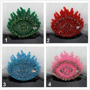 May include: Four eye-shaped beaded patches in green, red, blue, and pink. Each patch features a central pupil surrounded by concentric circles of beads and rhinestones, with radiating beaded eyelashes and a single teardrop-shaped crystal accent.