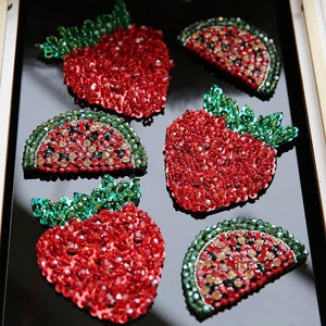 May include: Close-up of six fruit-shaped brooches. Three are strawberries, and three are watermelon slices. The brooches are covered in red, green, and clear sparkling crystals. The strawberries have green crystal leaves.
