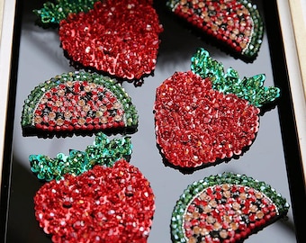 May include: Close-up of six fruit-shaped brooches. Three are strawberries, and three are watermelon slices. The brooches are covered in red, green, and clear sparkling crystals. The strawberries have green crystal leaves.