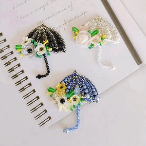 May include: Three beaded umbrella brooches with floral embellishments. The umbrellas are black, white, and blue. The flowers are white, yellow, and green.