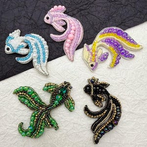 May include: Five colorful beaded fish ornaments. The fish are made of small beads in various colors, including pink, purple, yellow, green, and black. The fish are all different shapes and sizes, and they are all very detailed.