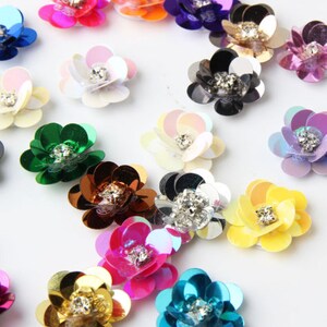 Puede incluir: Surtido de lentejuelas de colores en forma de flor con centros de pedrería. Las lentejuelas son iridiscentes y vienen en una variedad de colores, incluyendo rosa, azul, verde, amarillo y morado.