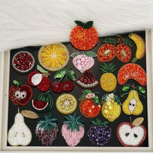 May include: A collection of assorted fruit-shaped patches crafted from colourful beads and sequins. The set includes depictions of oranges, lemons, strawberries, pears, cherries, pineapples, watermelon slices, and apples arranged on a black surface.