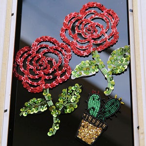 May include: Three embroidered patches featuring a cactus in a pot, and two roses with green leaves. The patches are made of sequins and beads in shades of red, green, and gold.