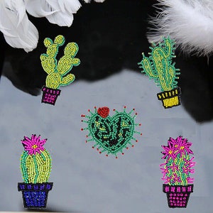 May include: Five colourful cactus-shaped beaded iron-on patches. The patches are green, yellow, pink, and red. The patches are in the shape of a heart, a traditional cactus, and a cactus with multiple arms.
