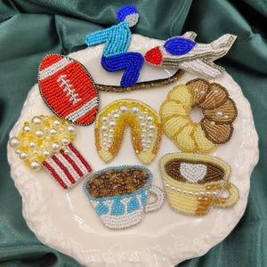 以下が含まれることがあります： 様々なデザインのカラフルなビーズ付きアイロン接着パッチのコレクション。フットボール、ポップコーン、ドーナツ、コーヒーカップ、馬蹄形、飛行機、青いジャンプスーツを着た人が含まれています。