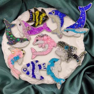 Puede incluir: Una colección de diez broches de animales hechos con cuentas que representan delfines, ballenas y peces. Los broches están hechos con cuentas y lentejuelas de colores, y son perfectos para añadir un toque de fantasía a cualquier atuendo.