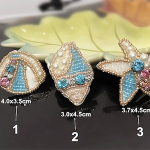 Puede incluir: Tres adornos de cuentas con forma de concha, concha y estrella de mar. Las conchas son blancas con detalles azules y rosas. La estrella de mar es blanca con detalles azules y rosas. Los adornos miden 4,0 x 3,5 cm, 3,0 x 4,5 cm y 3,7 x 4,5 cm.