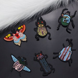 Puede incluir: Seis parches de hierro bordados que representan insectos coloridos y una mariposa. Los parches están hechos con lentejuelas y cuentas. Los insectos incluyen un escarabajo verde, un escarabajo amarillo y morado, un escarabajo azul y rosa, un escarabajo marrón y morado y una mosca azul y rosa.