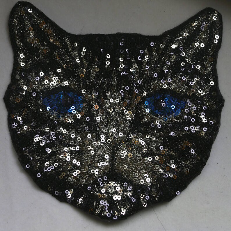 May include: A black cat face patch with silver and gold sequins and blue eyes. The patch is made of fabric and is perfect for adding a touch of whimsy to clothing or accessories.