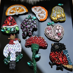 May include: Assortment of colourful beaded and sequined iron-on patches, including a watermelon, grapes, a pear, an apple, a carnation, a rooster, and two birds.