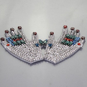 May include: A pair of silver-toned hand-shaped embellishments with rhinestones and colorful gemstones. The embellishments are joined at the fingertips and have a raised, textured design.