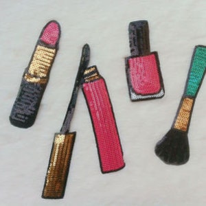 May include: Four colorful makeup items made of sequins, including a pink lipstick, a pink nail polish bottle, a black mascara wand, and a green makeup brush.