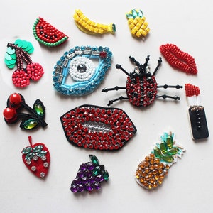 Puede incluir: Surtido de parches termoadhesivos coloridos con varios diseños, incluyendo una rodaja de sandía, un plátano, una piña, cerezas, una mariquita, unos labios, un mal de ojo, una uva, una fresa y un pintalabios.