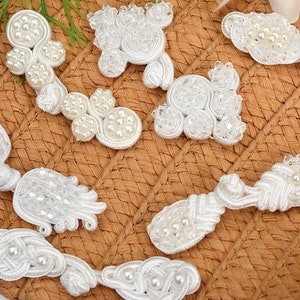 May include: Assortment of white pearl and fabric embellishments for sewing and crafting projects. The embellishments are in various shapes and sizes, including flowers, leaves, and knots.
