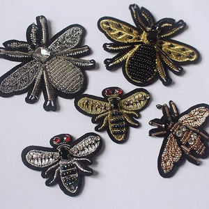 May include: Five embroidered iron-on patches featuring different insect designs. The patches are made with gold, silver, and black thread and have various embellishments, including beads and rhinestones. The insects include a bee, a dragonfly, and three different types of flies.