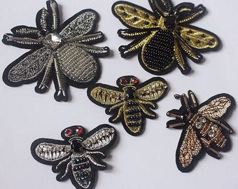 Könnte beinhalten: Fünf gestickte Bügelbilder mit verschiedenen Insektenmotiven. Die Patches sind aus goldenem, silbernem und schwarzem Garn gefertigt und haben verschiedene Verzierungen, darunter Perlen und Strasssteine. Die Insekten umfassen eine Biene, eine Libelle und drei verschiedene Arten von Fliegen.