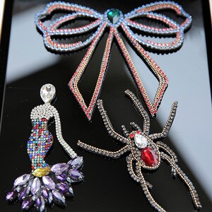 May include: Three jeweled appliques: a red, blue and gold bow, a purple and silver mermaid, and a black and red spider.