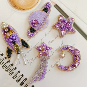 Könnte beinhalten: Satz von fünf lila und goldenen Perlenbroschen mit einer Rakete, einer Sternschnuppe, einem Halbmond, einem Stern und einem Planeten. Die Broschen sind mit Perlen und Strasssteinen verziert.