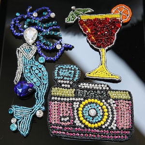 May include: A set of three colorful beaded patches featuring a mermaid, a camera, and a cocktail. The mermaid is blue and silver, the camera is pink, yellow, and black, and the cocktail is red, yellow, and orange.