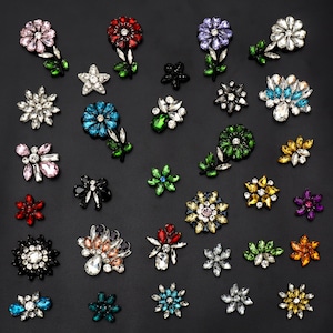 May include: Assortment of colorful rhinestone flower embellishments with various designs and colors, including red, blue, green, purple, and black.