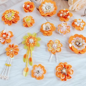 Puede incluir: Surtido de flores de tela naranja y amarilla con detalles de perlas. Las flores están dispuestas sobre una superficie blanca.