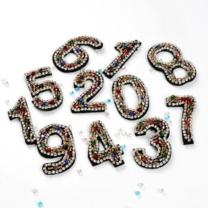 May include: Ten colorful, jeweled, iron-on patches in the shape of numbers 1 through 9 and 0. The patches are decorated with colorful rhinestones and beads.