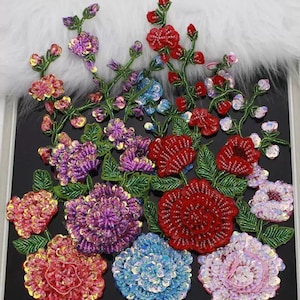 Puede incluir: Una colección de apliques florales coloridos con lentejuelas. Los diseños incluyen flores rojas, moradas, rosas y azules con hojas y tallos verdes. Los apliques están dispuestos sobre un fondo negro.