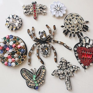 May include: Assortment of colourful beaded iron-on patches, including a spider, butterfly, dragonfly, heart, cross, and flower. The patches are made with rhinestones, beads, and sequins.