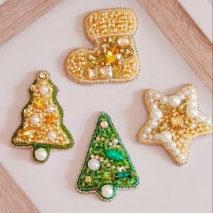 May include: Four Christmas-themed brooches, each featuring a different design: a stocking, a star, and two Christmas trees. The stocking is cream-coloured with gold and clear rhinestones. The star is cream-coloured with white pearls and gold rhinestones. One Christmas tree is green with green and clear rhinestones, and the other is cream-coloured with gold and clear rhinestones.