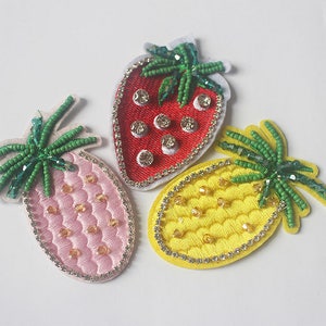 Puede incluir: Tres parches de frutas bordadas: una fresa roja, una piña rosa y una piña amarilla. Cada parche presenta detalles de cuentas, bordes de pedrería y copas de cuentas verdes. La fresa tiene pedrería transparente.