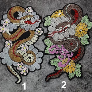 Puede incluir: Dos parches de planchado bordados con un diseño de serpiente con acentos florales. La serpiente de la izquierda es marrón y roja, mientras que la serpiente de la derecha es marrón y negra. Ambos parches tienen un fondo blanco con flores rosas, amarillas y verdes.
