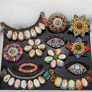 Könnte beinhalten: Sortiment an bunten Perlen- und Muschelverzierungen. Die Verzierungen zeigen verschiedene Designs, darunter Blumen, Augen und geometrische Muster. Die Verzierungen sind perfekt, um Kleidung, Taschen und anderen Accessoires einen Hauch von handgemachtem Charme zu verleihen.
