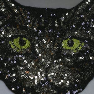 May include: Black sequin cat face applique with green eyes. The cat face is made of sequins and has a black background.
