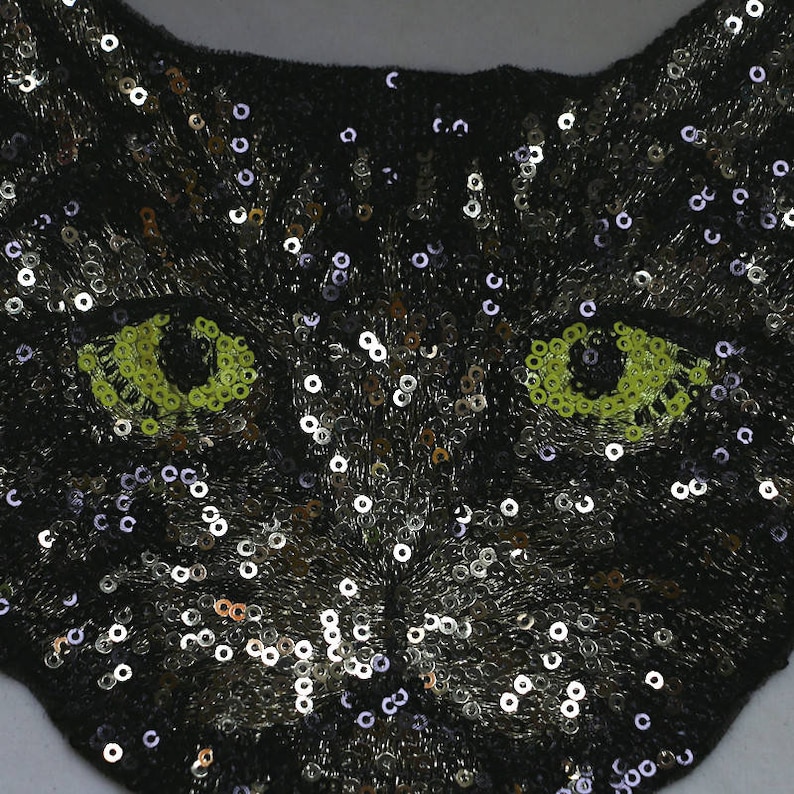 May include: Black sequin cat face applique with green eyes. The cat face is made of sequins and has a black background.