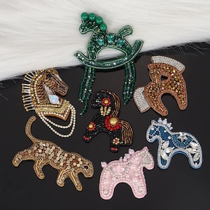 Puede incluir: Una colección de broches con forma de caballo y animales con cuentas de varios colores. Los broches incluyen un caballito balancín verde, un caballo marrón con crin blanca y un caballo rosa con detalles de perlas. Otros broches incluyen un tigre y un caballo azul.