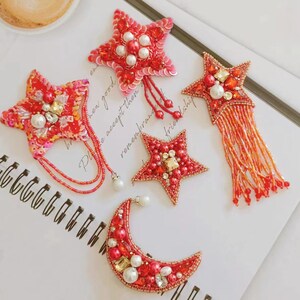 May include: Five red and gold beaded star and crescent moon shapes with pearl accents. The stars have dangling beaded strands. The crescent moon has a beaded fringe.