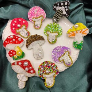 May include: Assortment of colourful beaded mushroom-shaped brooches. The brooches feature various colours, including red, green, purple, yellow, and pink. Each brooch has a pearl-like bead in the centre of the mushroom cap.