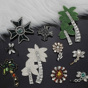 May include: A collection of decorative beaded and jeweled appliques in various shapes, including flowers, palm trees, and crosses. The appliques feature a variety of colors, including green, silver, black, and gold, and are embellished with rhinestones and beads.