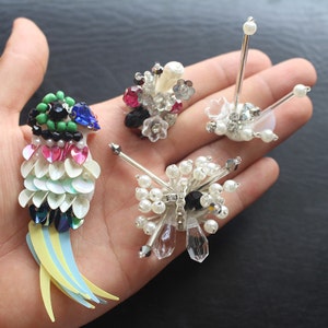 May include: Four colorful beaded brooches, including a parrot design with a blue head, green body, and yellow and blue tail feathers, a white and silver design with a black center, a white and silver design with a black center, and a pink, white, and silver design with a black center.