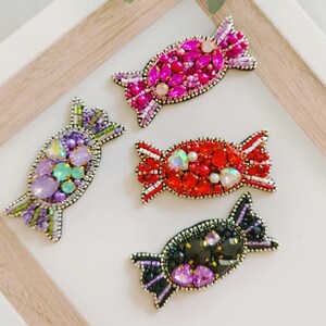May include: Four colourful candy-shaped brooches with beaded embellishments. The brooches are in shades of pink, purple, red, and black.
