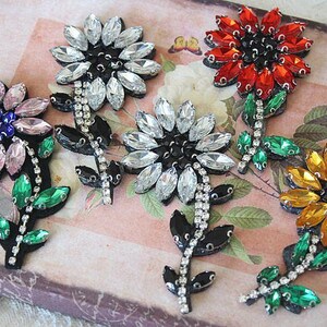 May include: Five rhinestone flower appliques with black centres and green leaves. The flowers are in various colours, including pink, white, red, and gold.
