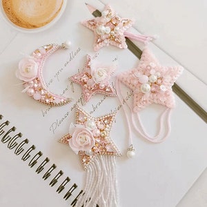 Könnte beinhalten: Fünf rosa und weiße gestickte Verzierungen mit Perlenakzenten. Die Verzierungen umfassen einen Halbmond, einen Stern mit Quaste und drei Sterne mit Perlenketten.
