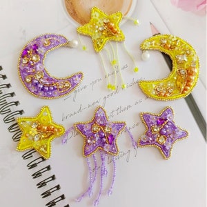 May include: Five embroidered patches featuring stars and crescent moons. The patches are made with beads, sequins, and pearls in shades of yellow, purple, and gold. The patches have a dangling fringe of beads.