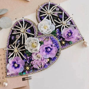 May include: Two heart-shaped brooches with intricate floral designs. The brooches feature black and purple backgrounds, adorned with white, lavender, and purple fabric flowers, beads, and sequins. Each brooch has a pearl drop. The brooches are handmade.