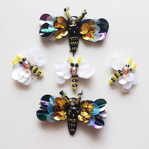 May include: Five colorful embroidered butterfly appliques. Two are white with yellow and black accents, two are iridescent blue, gold, and purple, and one is iridescent green, gold, and purple.